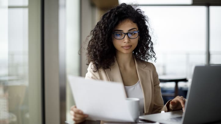 A woman in glasses is working on a laptop in an office.