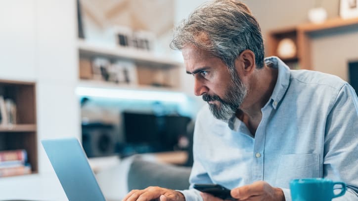 A man with a beard is using a laptop and cell phone.