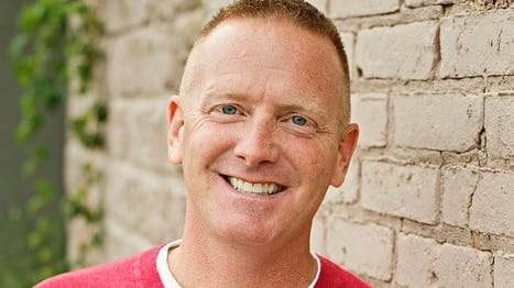 A man in a red v - neck shirt leaning against a brick wall.