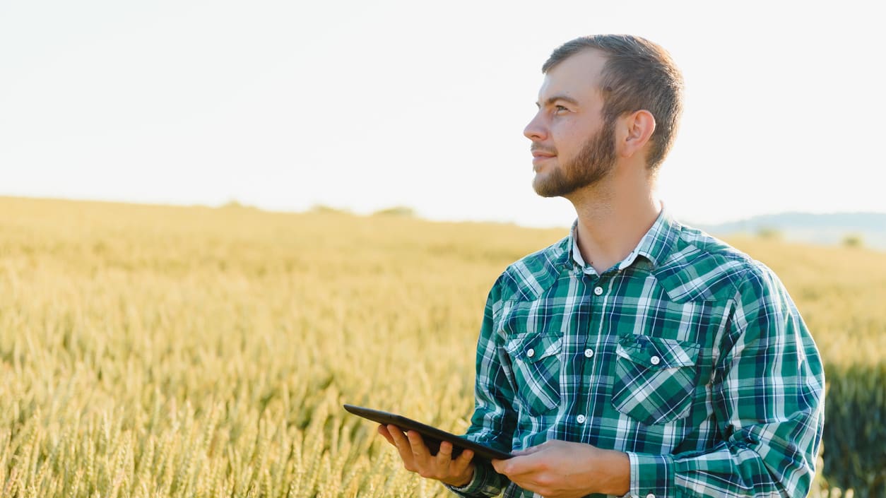 A man in a plaid shirt is standing in a wheat field with a tablet computer.