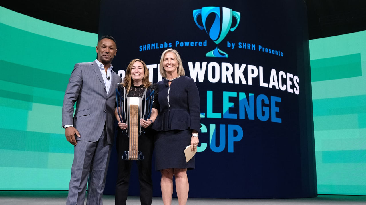 Three people standing on stage with a trophy for the best workplaces challenge.