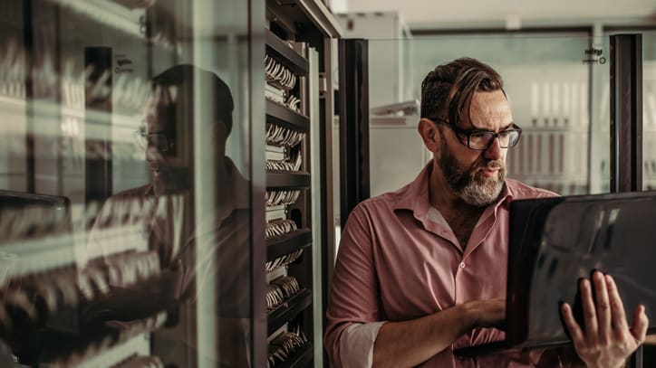 A man in glasses is using a laptop in a server room.