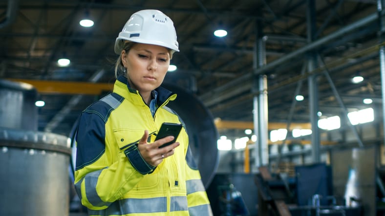 A woman in a hard hat is using a cell phone in a factory.