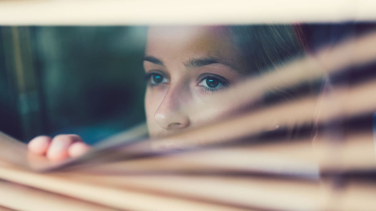 A woman looking out of a window with blinds.