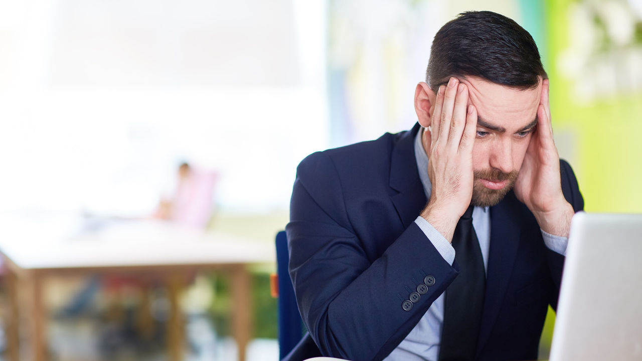 A man in a suit is holding his head in front of his laptop.