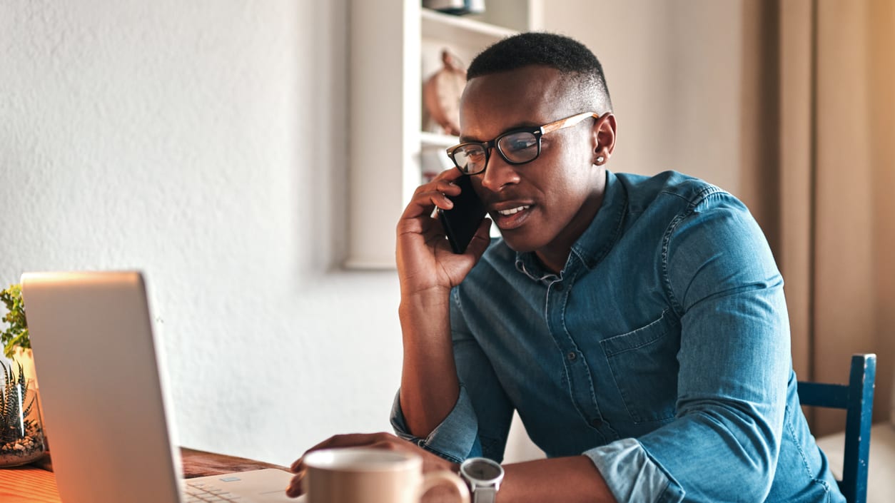 A man is talking on the phone while sitting at a desk with a laptop.