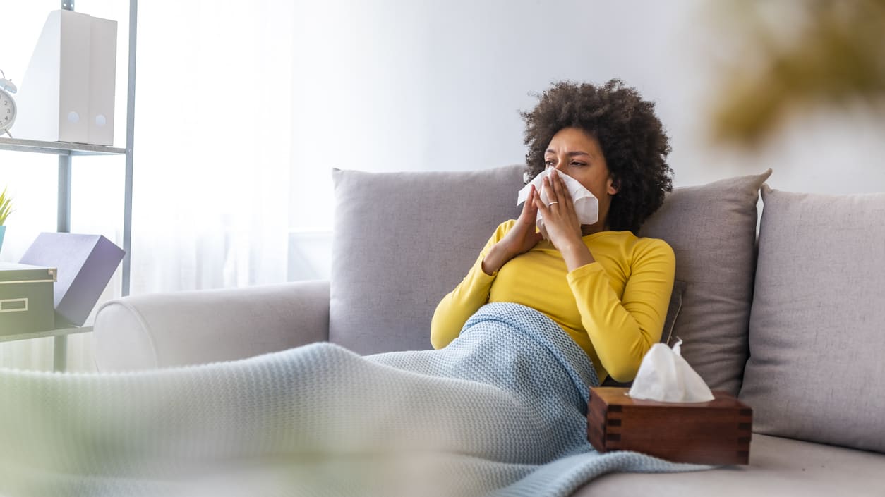 A woman with a cold lying on a couch and blowing her nose.