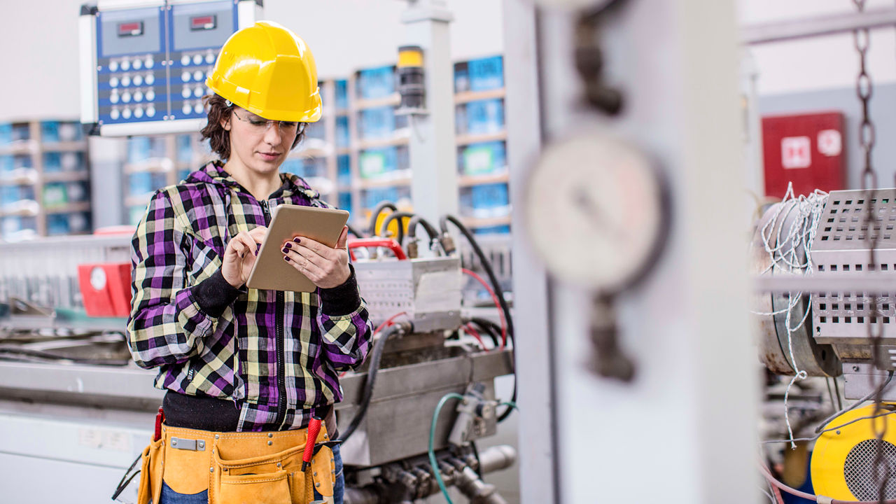 A woman in a hard hat is using a tablet in a factory.