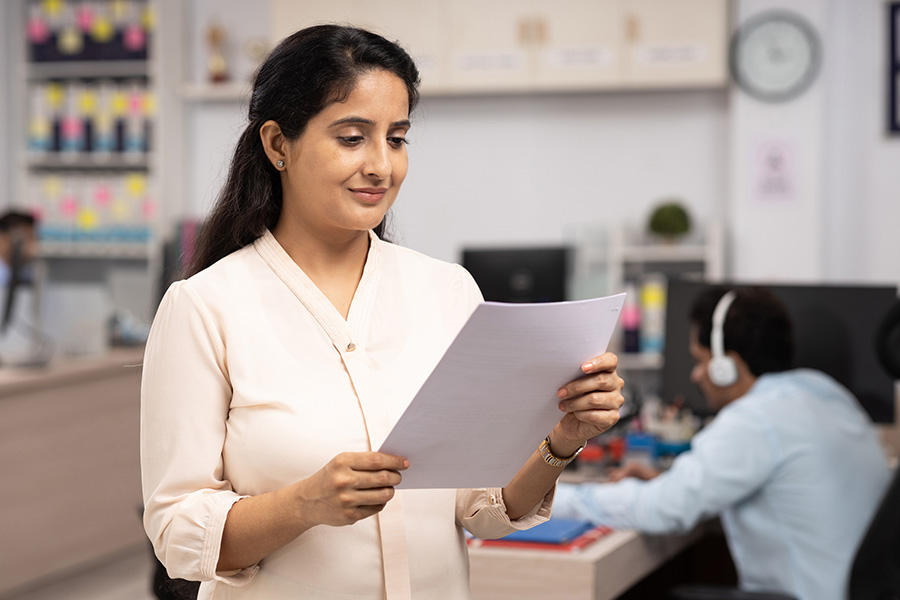 A business professional woman holding a piece of paper with someone behind her at his cubicle with headphones on. and a clock in the background of the office setting.