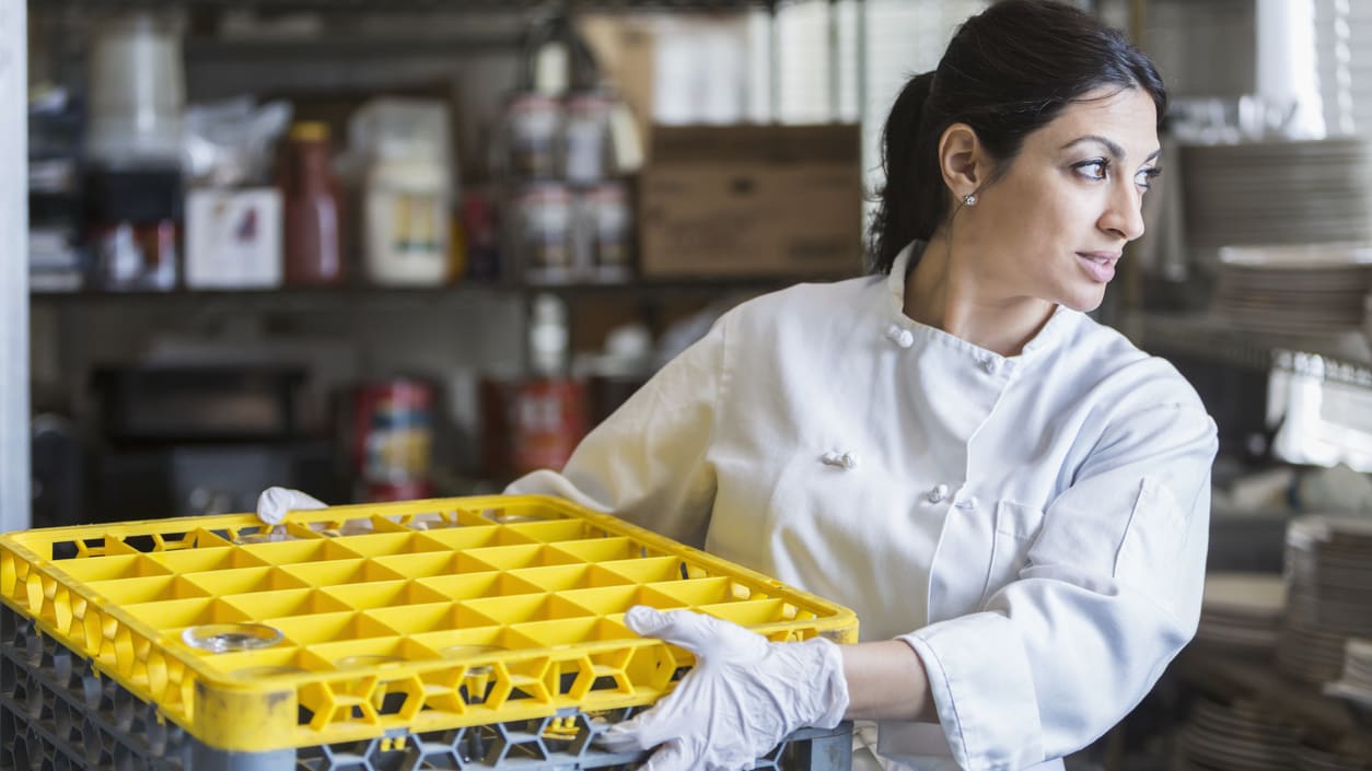 A female chef holding a yellow crate in a kitchen.