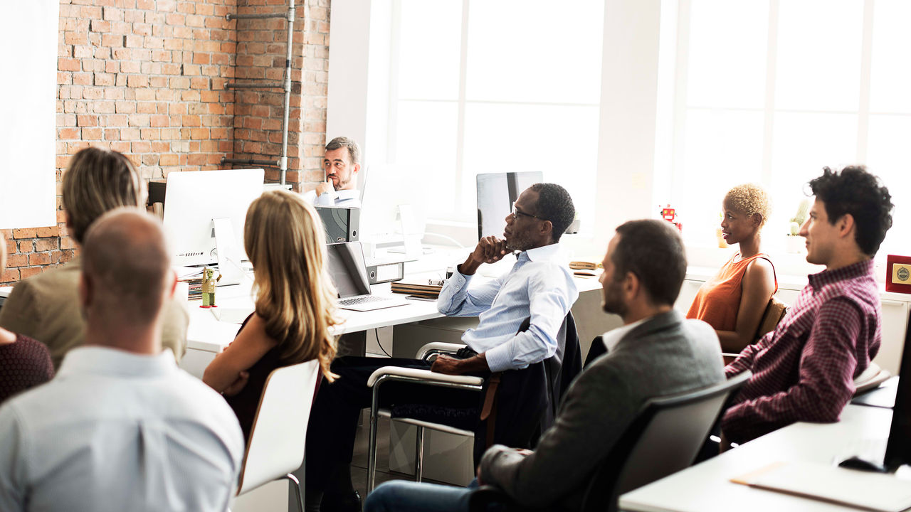 A group of people sitting at a desk in front of a white board.