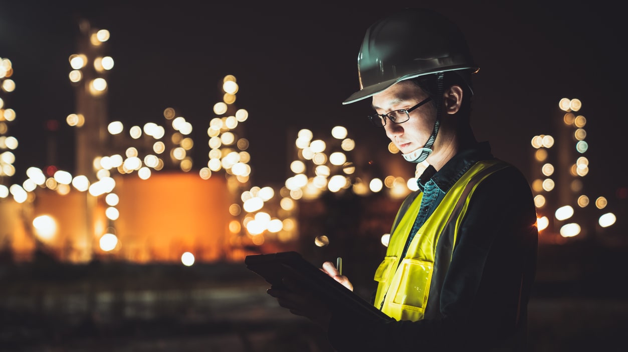 A worker using a tablet in front of an oil refinery at night.