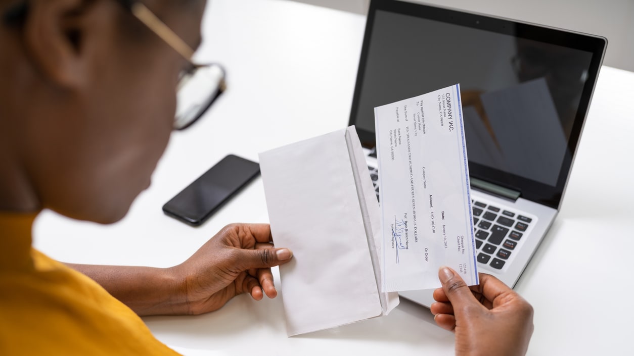 A woman is holding a check in front of a laptop.