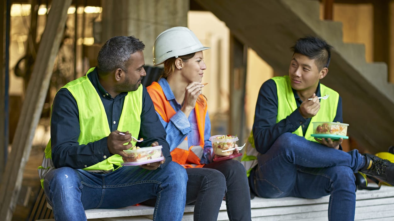 Three construction workers sitting on a bench eating food.