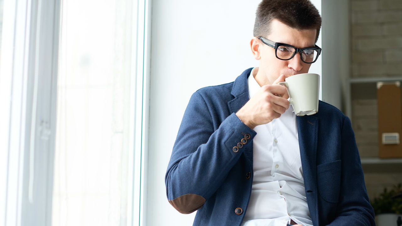 A man in glasses is sitting on a window sill and drinking a cup of coffee.
