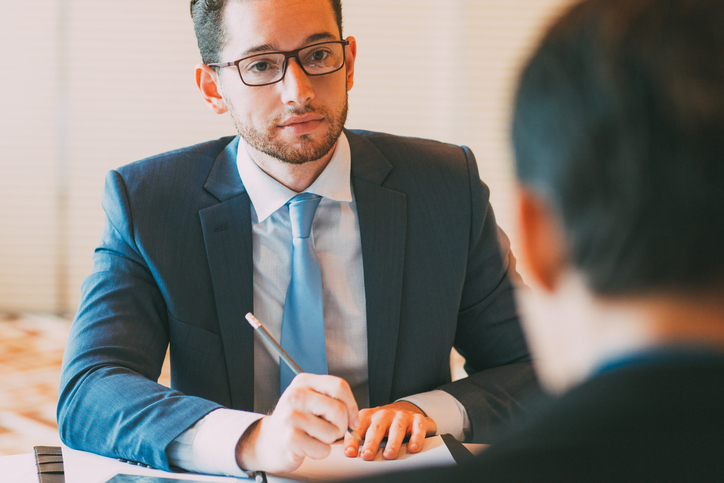 An interviewer with a pencil poised above a sheet of paper across the desk from a job applicant