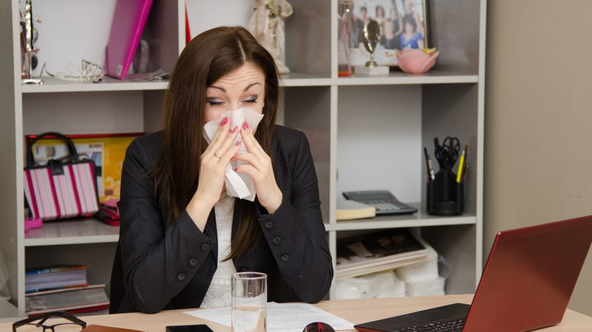 A woman in a business suit is blowing her nose at her desk.