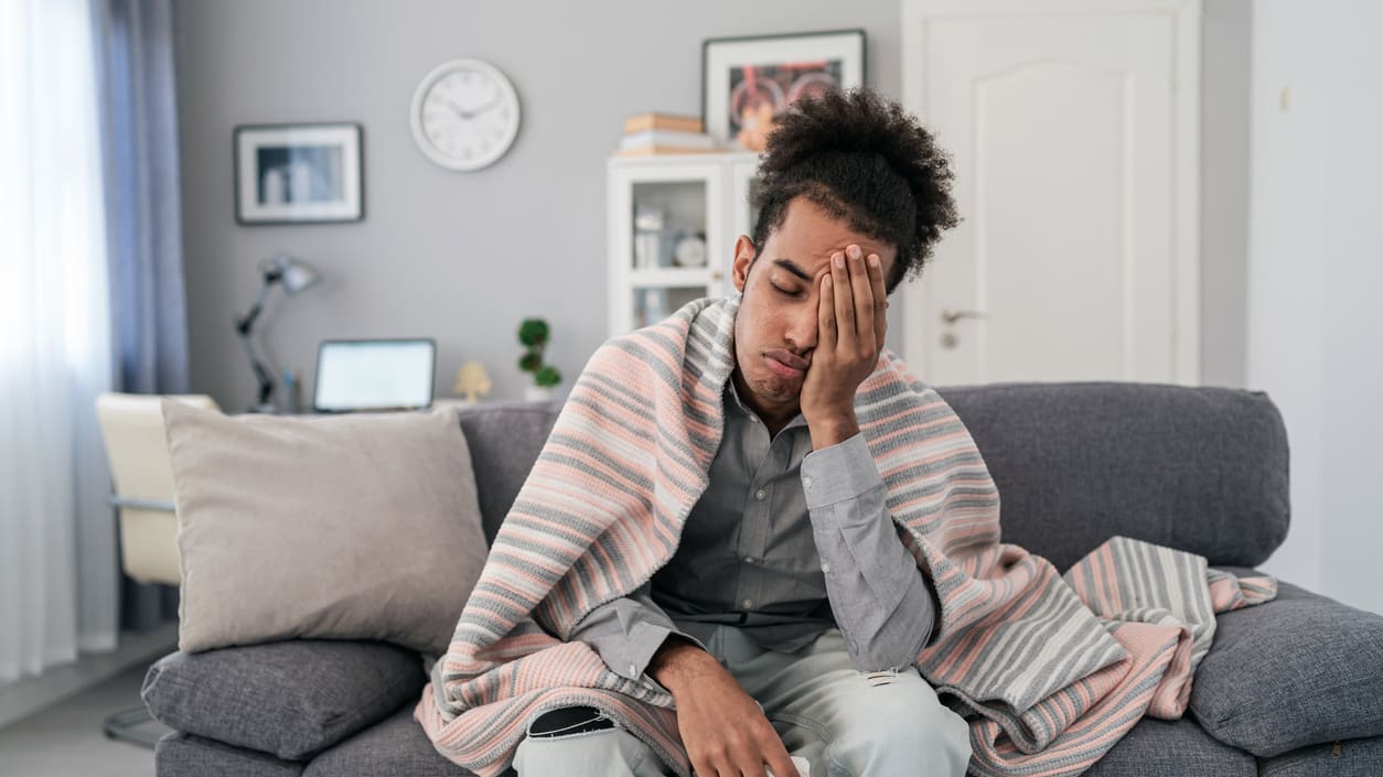 A man sitting on a couch with a blanket on his shoulders holding his head