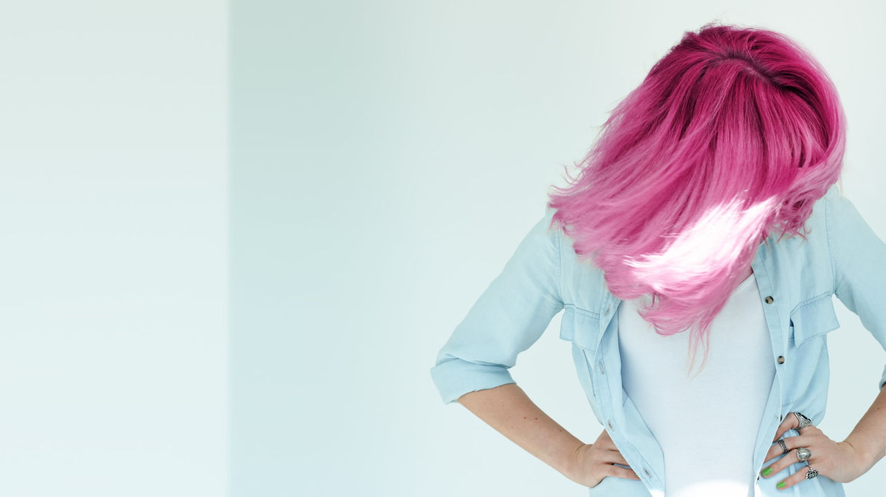 A woman with pink hair is standing in front of a white wall.