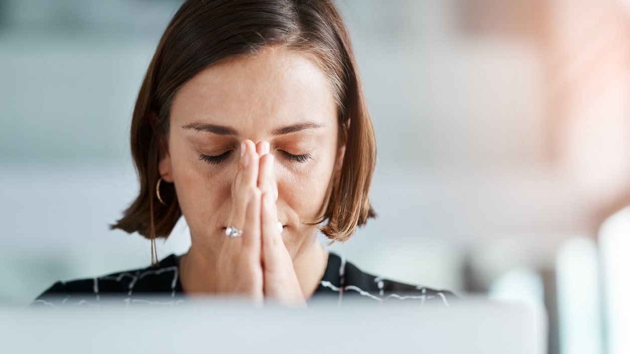 A woman with her hands covering her face while working on a laptop.