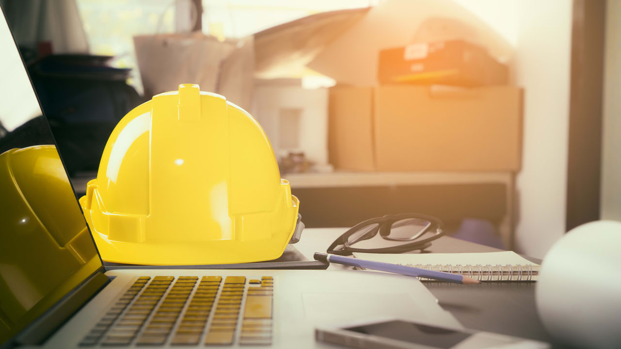 A yellow hard hat sits on a desk next to a laptop.