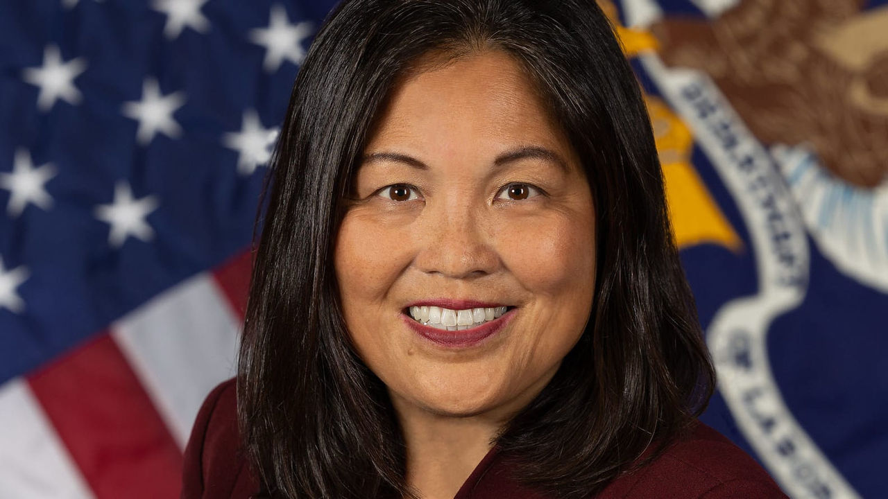 A woman in a maroon jacket standing in front of an american flag.