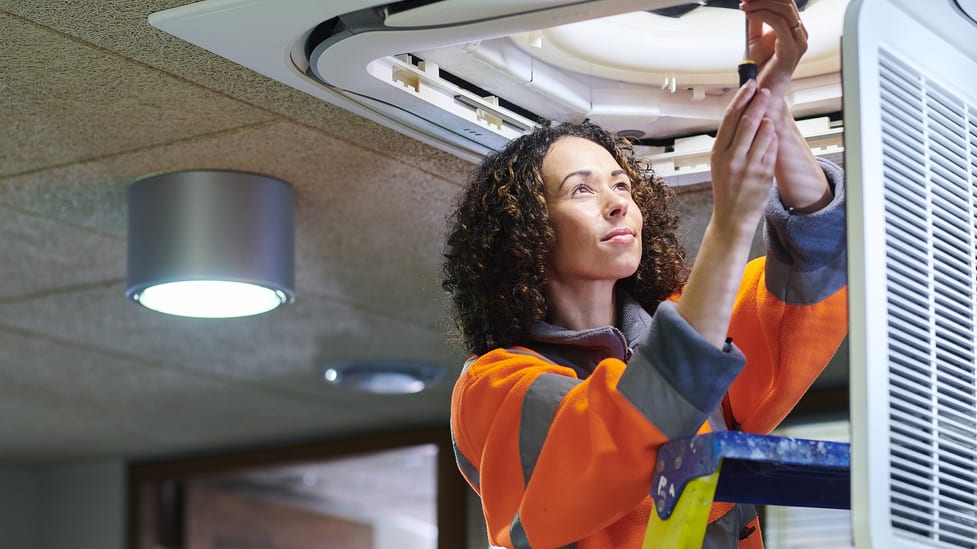 A woman on a ladder working on an air conditioning unit.