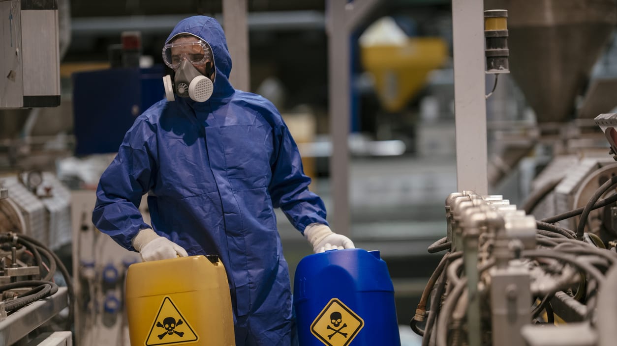 A worker in a protective suit is holding a bucket of chemicals.