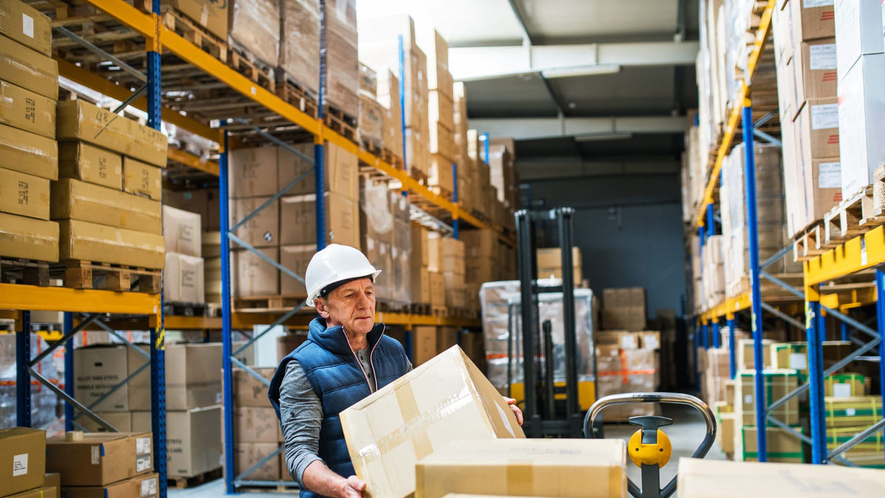A man in a hard hat is holding boxes in a warehouse.