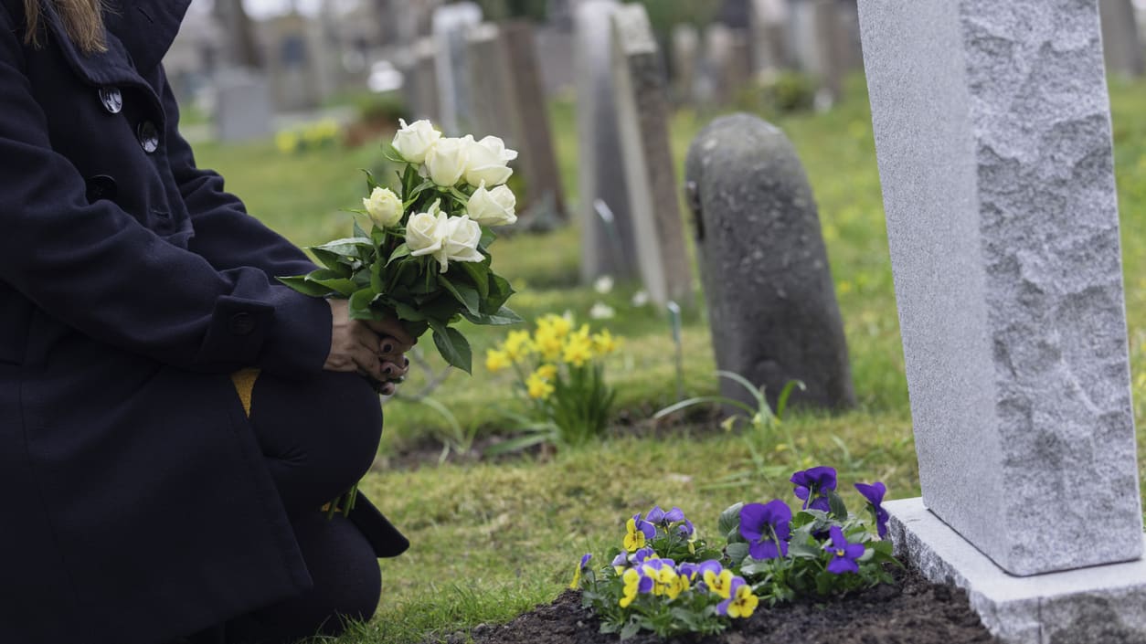 A woman kneeling down next to a grave with flowers.