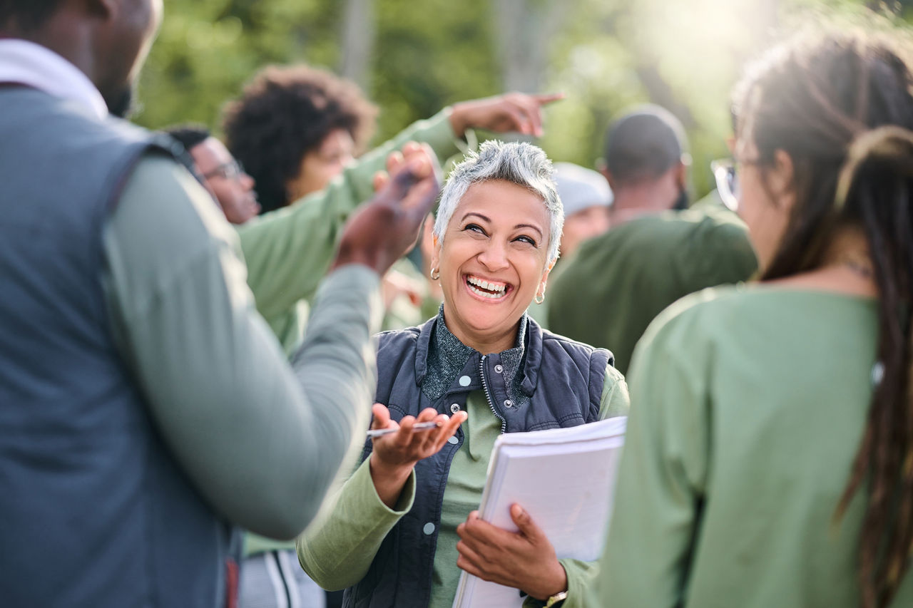 A smiling woman with short gray hair engages animatedly with a diverse group of people outdoors. She's holding papers and appears to be in mid-conversation, possibly at a casual outdoor meeting or community event.