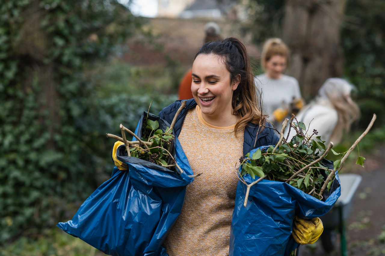 A smiling woman in a blue jacket and mustard sweater participates in a community garden clean-up, holding two blue bags filled with garden waste. In the background, other volunteers are also engaged in gardening activities, contributing to the community effort.