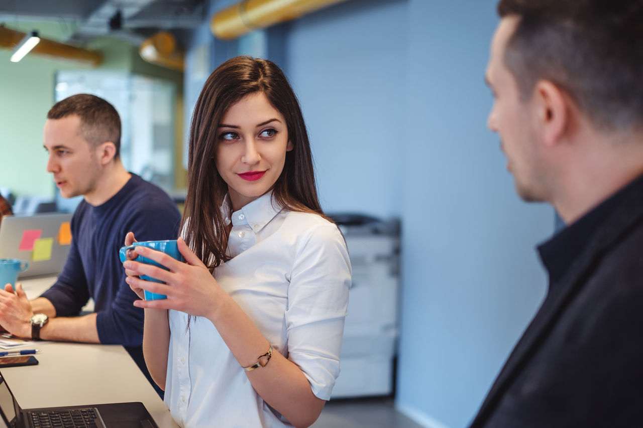 A professional young woman in a white shirt holds a blue mug, giving a side glance towards a male colleague while in a modern office setting with another coworker in the background focused on his laptop.