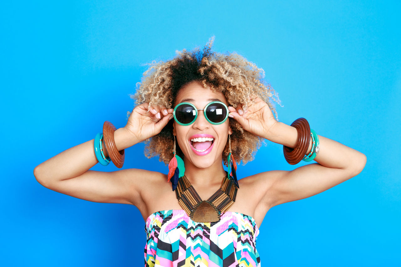 A joyful woman with curly hair wearing summer fashion accessories—sunglasses on her eyes, various bangles on her wrist, and feather earrings—strikes a playful pose against a vibrant blue background.