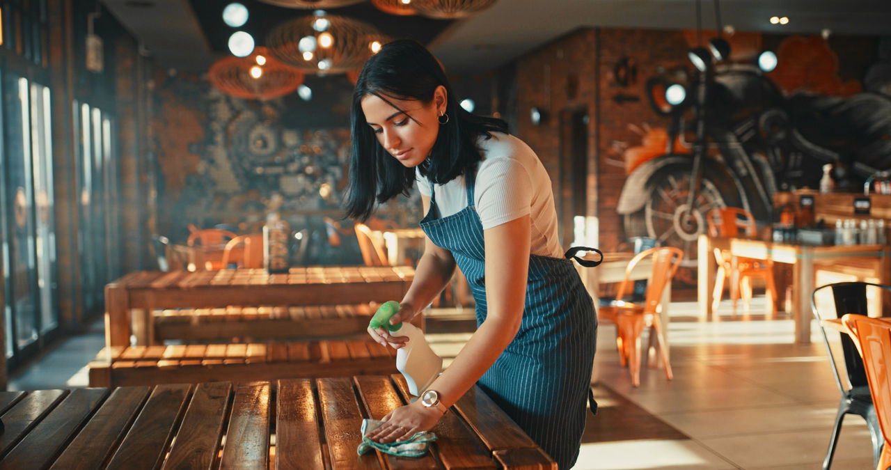 A young woman, wearing a blue-striped apron over a white shirt, is cleaning a wooden table in a warmly lit restaurant. Her focused expression and diligent work reflect the demanding nature of front-line service jobs.