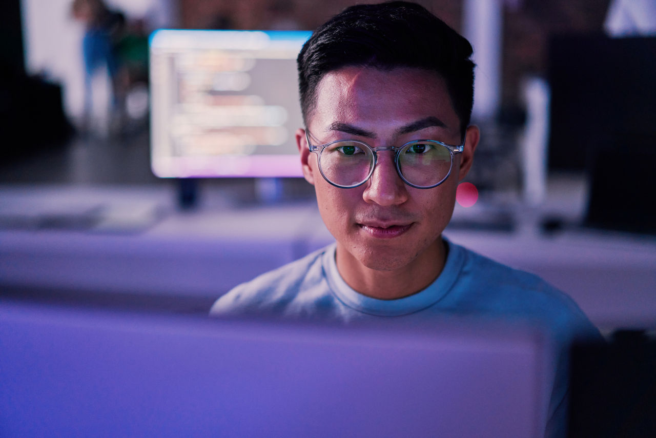 A young man with glasses is lit by the soft glow of a computer screen, hinting at late hours in a tech environment. He's intently focused on his work, reflecting the concentration and dedication common in the tech industry.