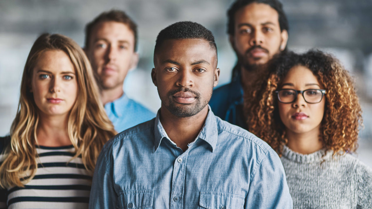 A diverse group of five young professionals standing in a line, with a focused man in the foreground. They represent a cross-section of the workforce, with varied expressions reflecting different levels of engagement and confidence.
