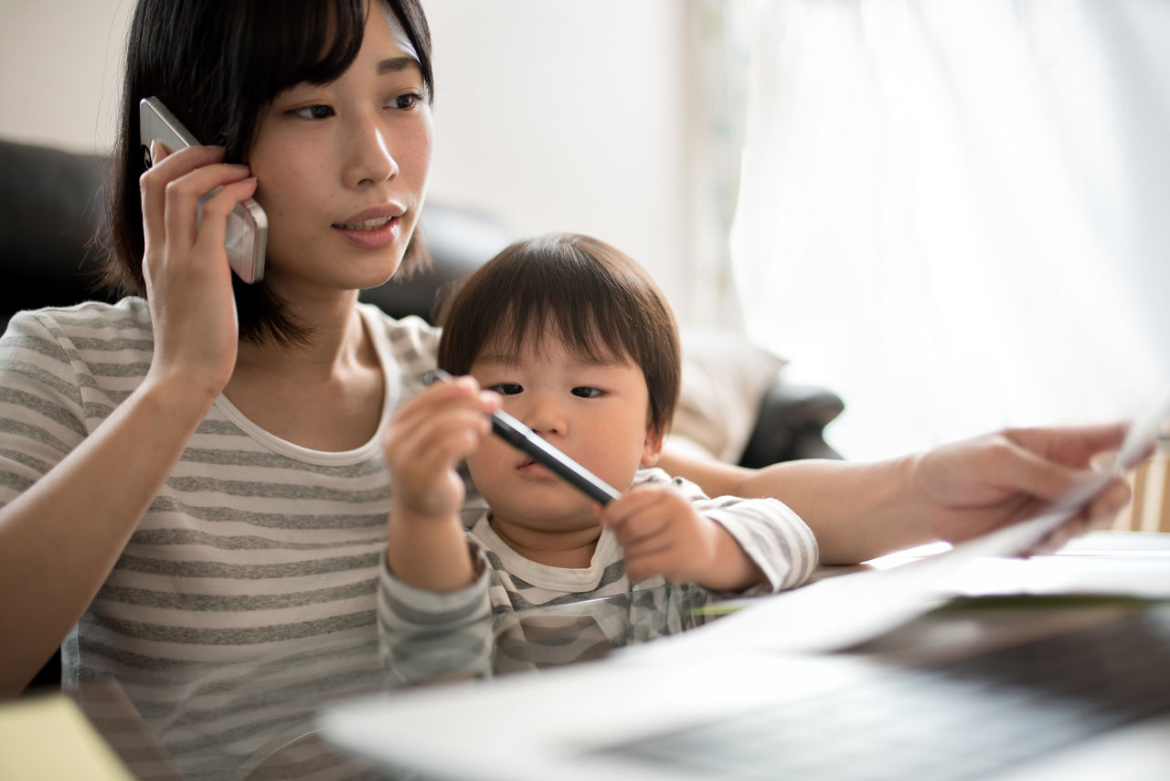 A young Asian woman multitasking at home, talking on a cell phone while her toddler, who's sitting in her lap, plays with a pen. She's reviewing documents spread out in front of her, symbolizing the balance of work and family life.