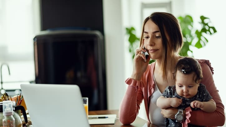A woman talking on the phone in front of a laptop while holding a baby