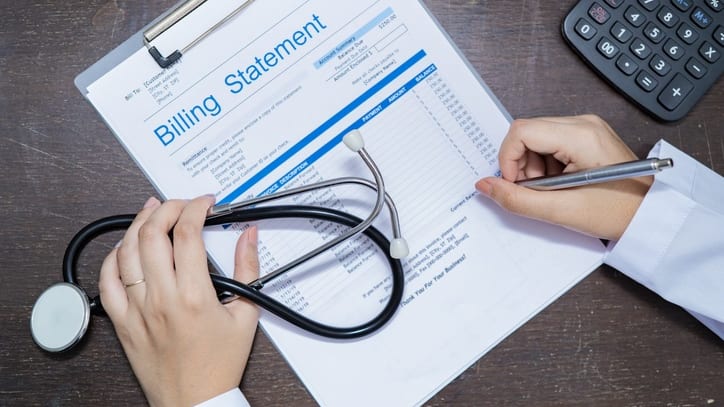 A doctor's hand holding a stethoscope and writing a billing statement on a clipboard.