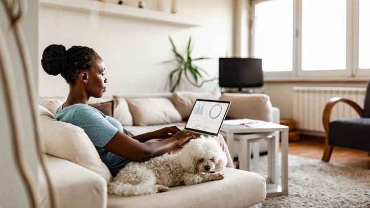 A woman using a laptop while sitting on a couch with her dog.