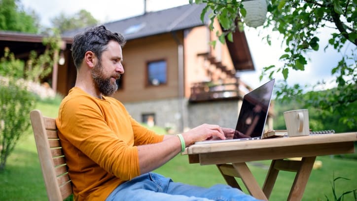 A bearded man sitting on a wooden bench using a laptop.