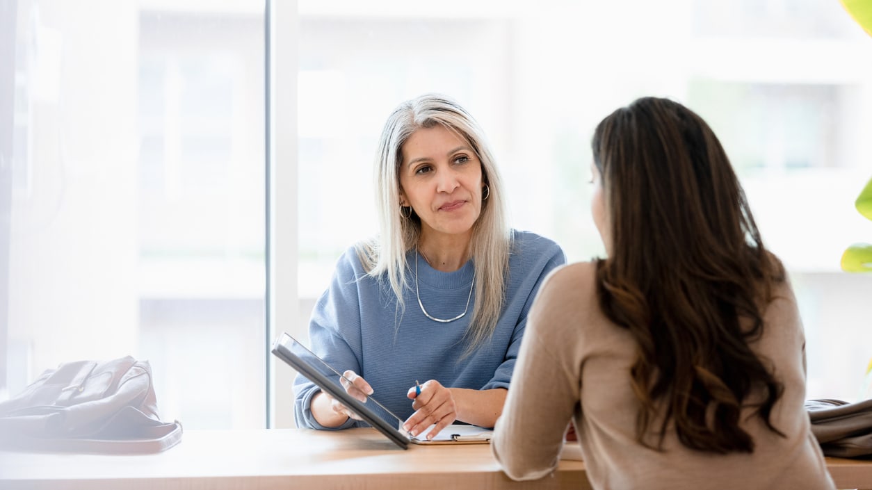 Two women sitting at a desk talking to each other.