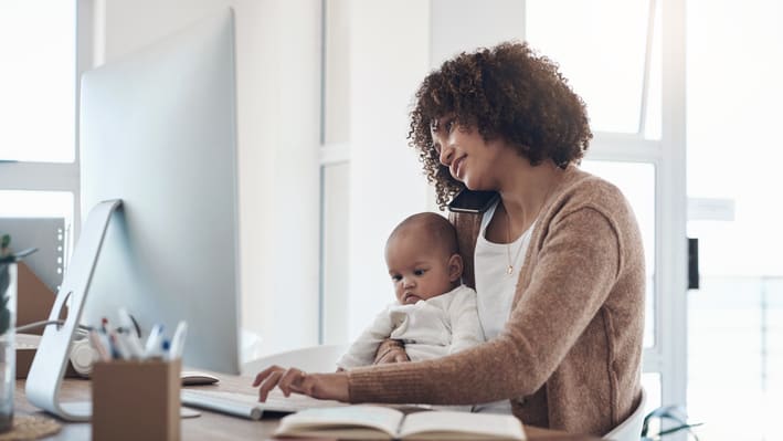 A woman holding a baby while working at her computer.