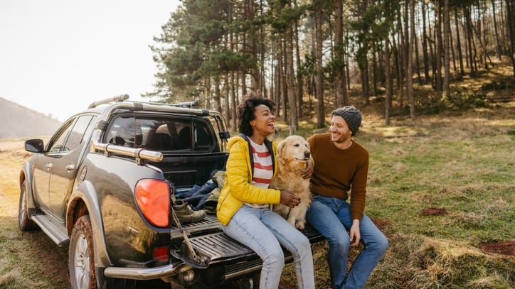 A couple sits in the back of a pickup truck with their dog.