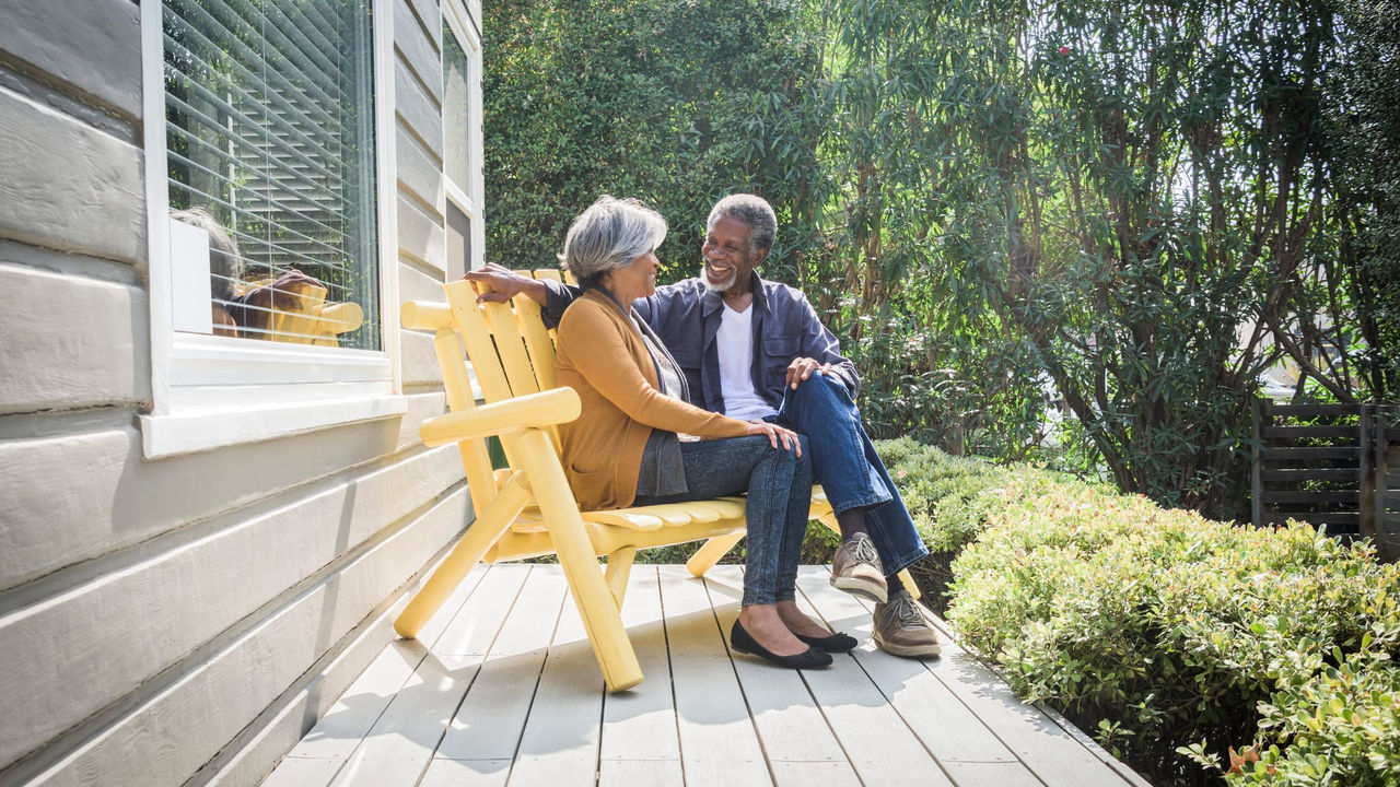 An older couple sitting on a yellow chair outside their home.