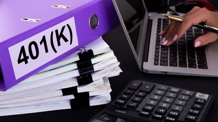 A person is typing on a laptop while holding a purple folder with the word 401k on it.