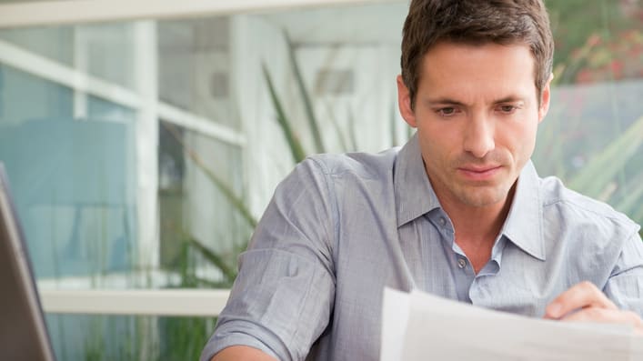 A man looking at a piece of paper in front of a laptop.