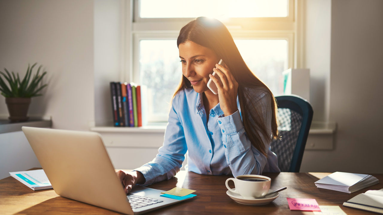 A woman is talking on the phone while sitting at a desk with a laptop.