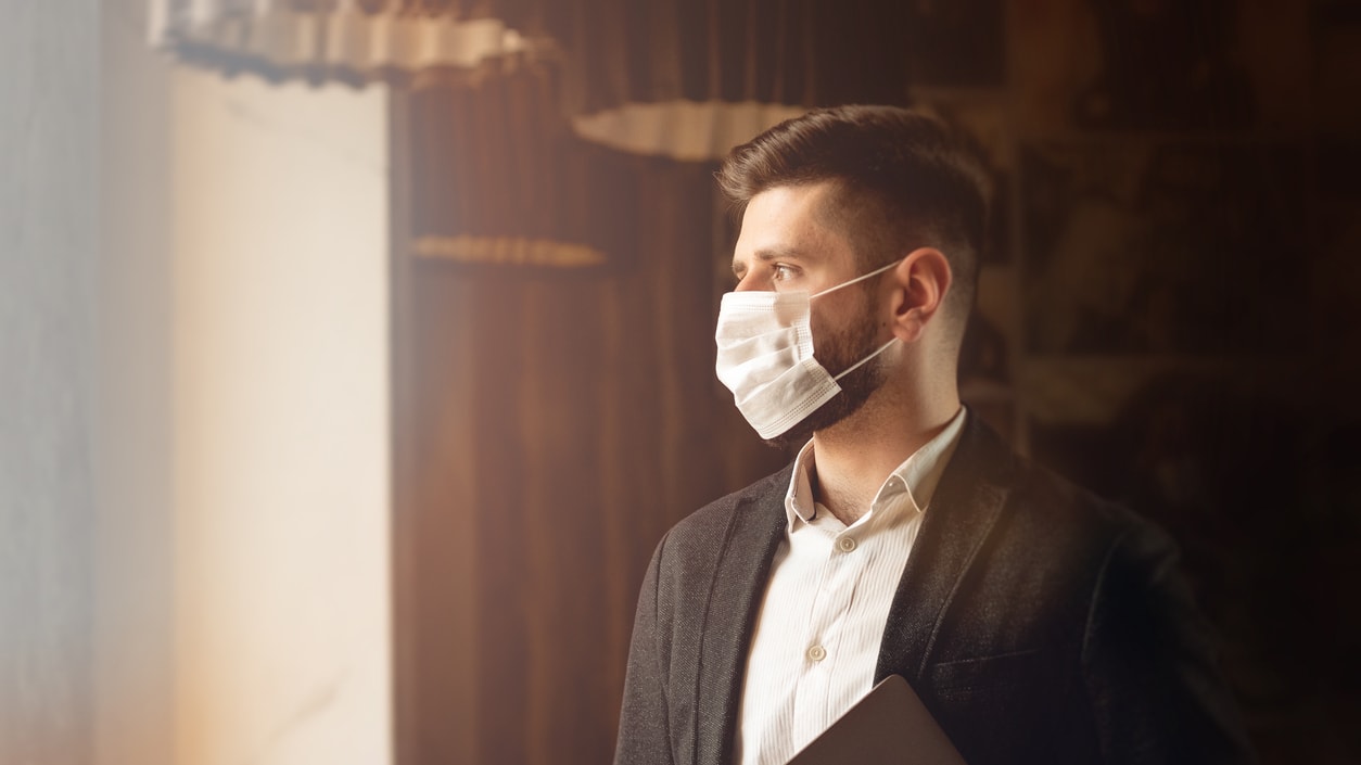 A businessman wearing a surgical mask in front of a window.